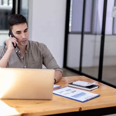 man in gray shirt sitting at a laptop talking on the phone