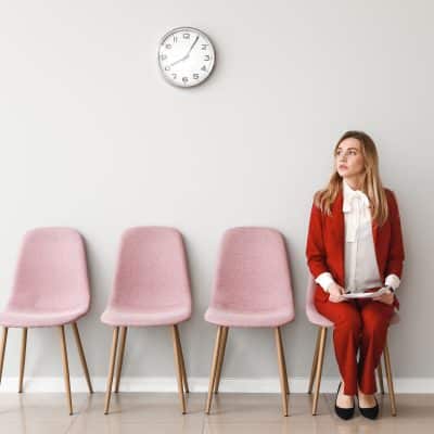woman in red suit with papers on her lap sitting in a row of pink chairs