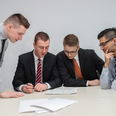 4 men in suits gathered together looking at papers on a conference table